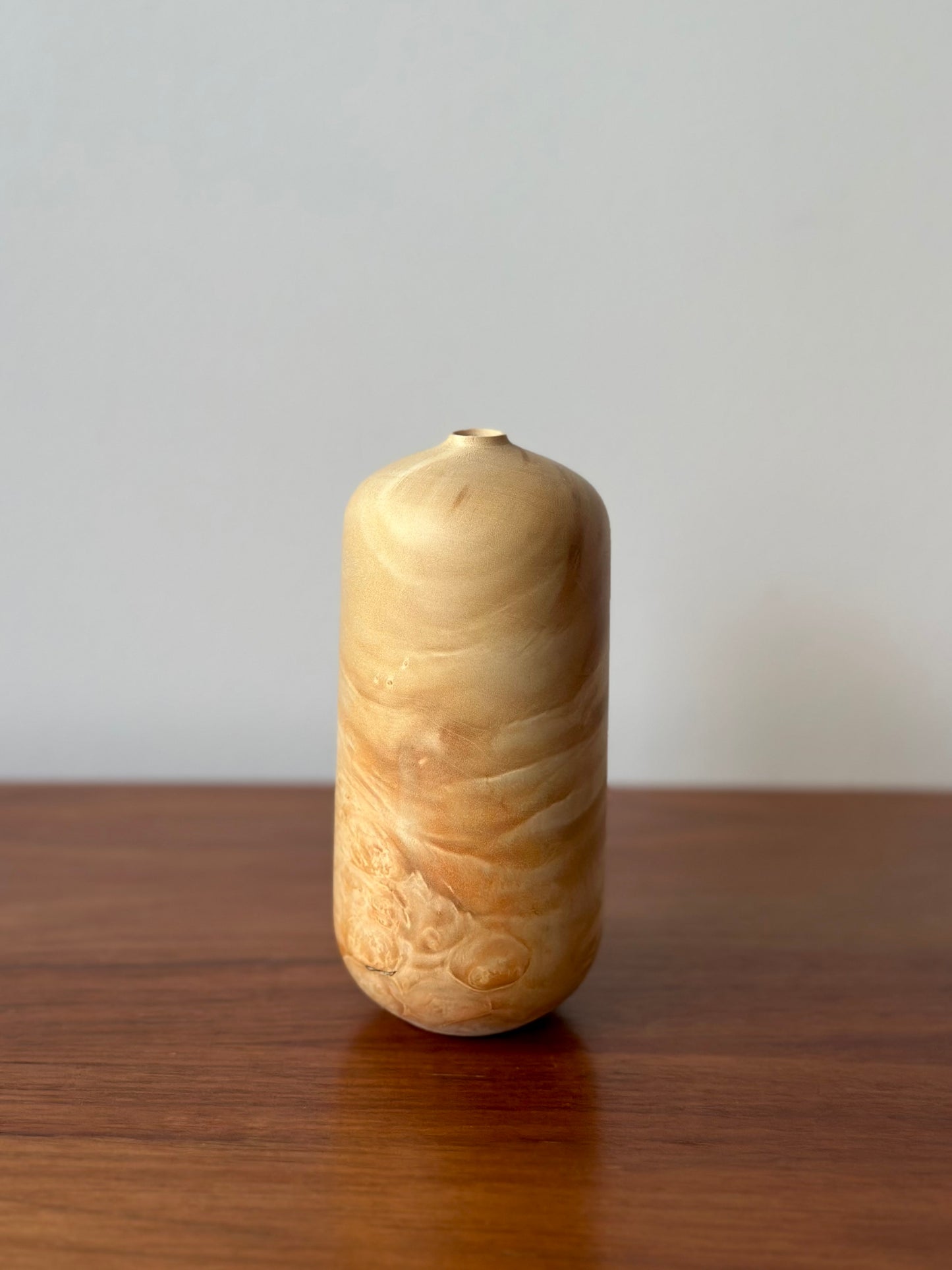 A small cylindrical ceramic vase with a rounded body and narrow neck, featuring a marbled swirl pattern in light beige, cream, and soft tan tones. The vase is standing on a polished dark brown wooden surface against a plain light gray wall.