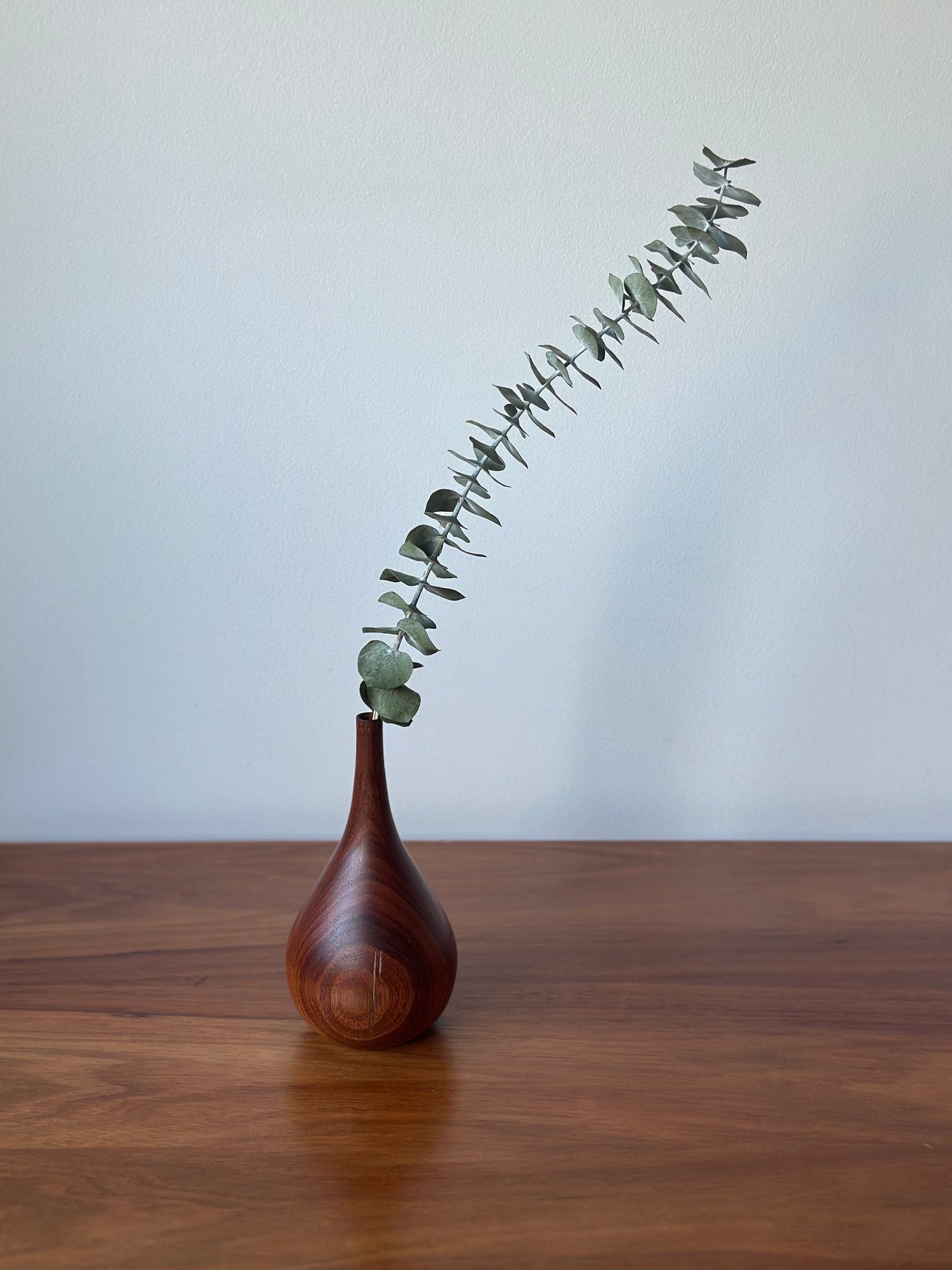 A small teardrop-shaped wooden vase with a glossy finish, holding a single eucalyptus stem, set on a wooden tabletop against a plain light gray wall.
