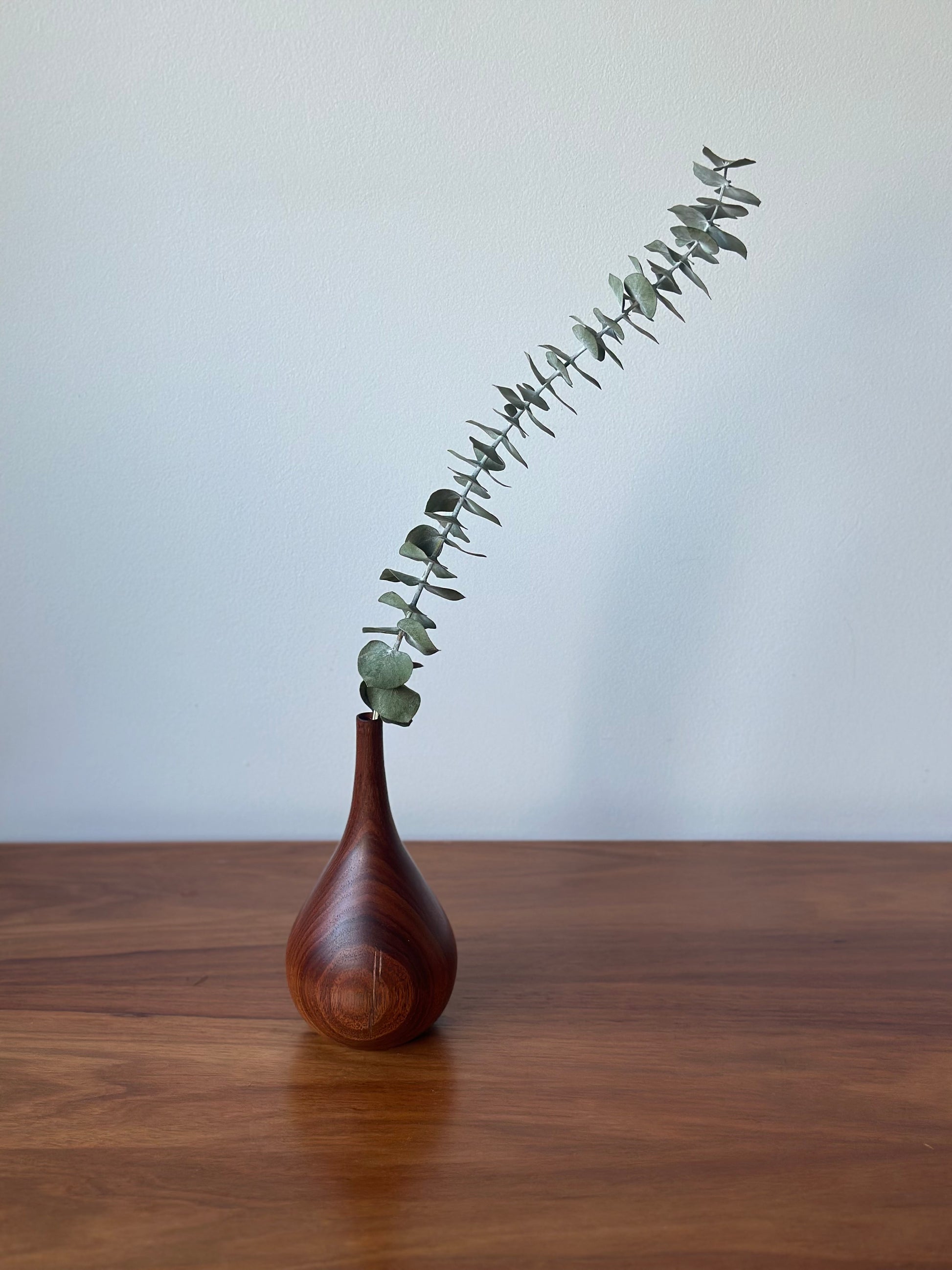 A small teardrop-shaped wooden vase with a glossy finish, holding a single eucalyptus stem, set on a wooden tabletop against a plain light gray wall.