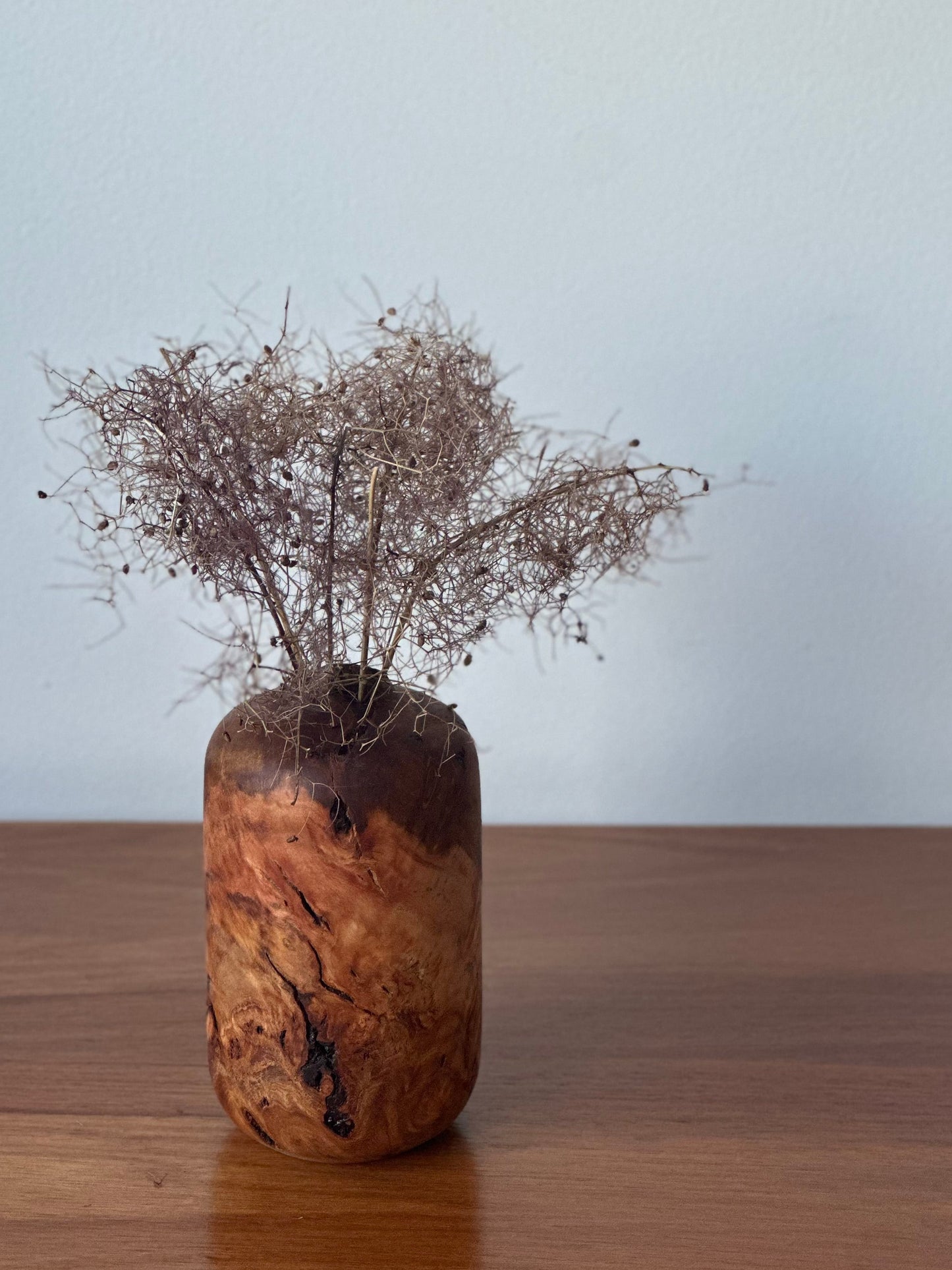 A small cylindrical vase made from natural burl wood with a dark brown top and warm tan-brown marbled body, holding dried brown grass stems, set on a wooden tabletop against a plain light gray wall.