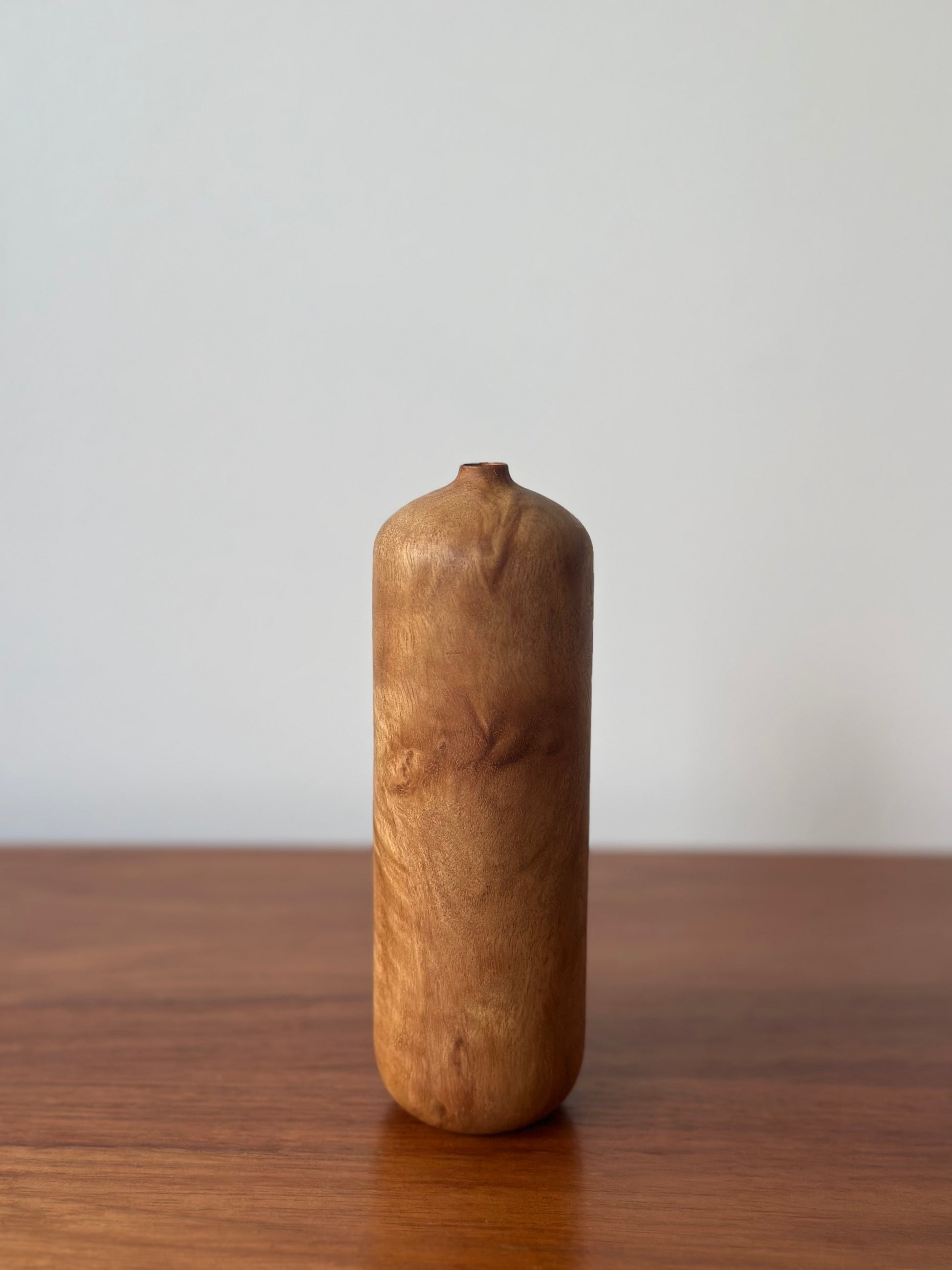 A small cylindrical wooden vase with a natural burl wood grain pattern, standing on a wooden table against a plain light gray wall.