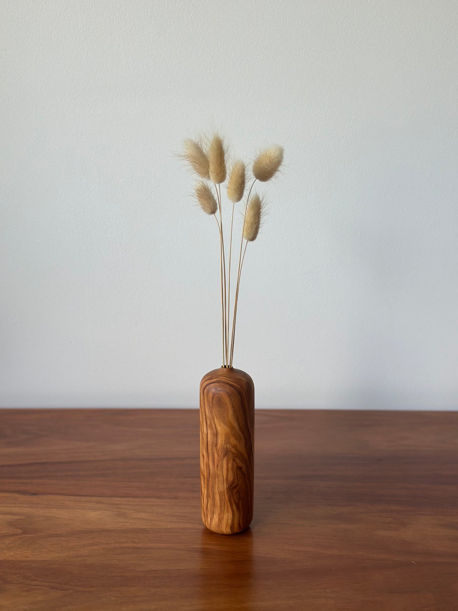 A small cylindrical wooden vase with a natural grain pattern, holding dried bunny tail grass stems, set on a wooden tabletop against a plain light gray wall.