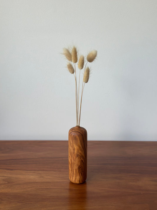 A small cylindrical wooden vase with a natural grain pattern, holding dried bunny tail grass stems, set on a wooden tabletop against a plain light gray wall.