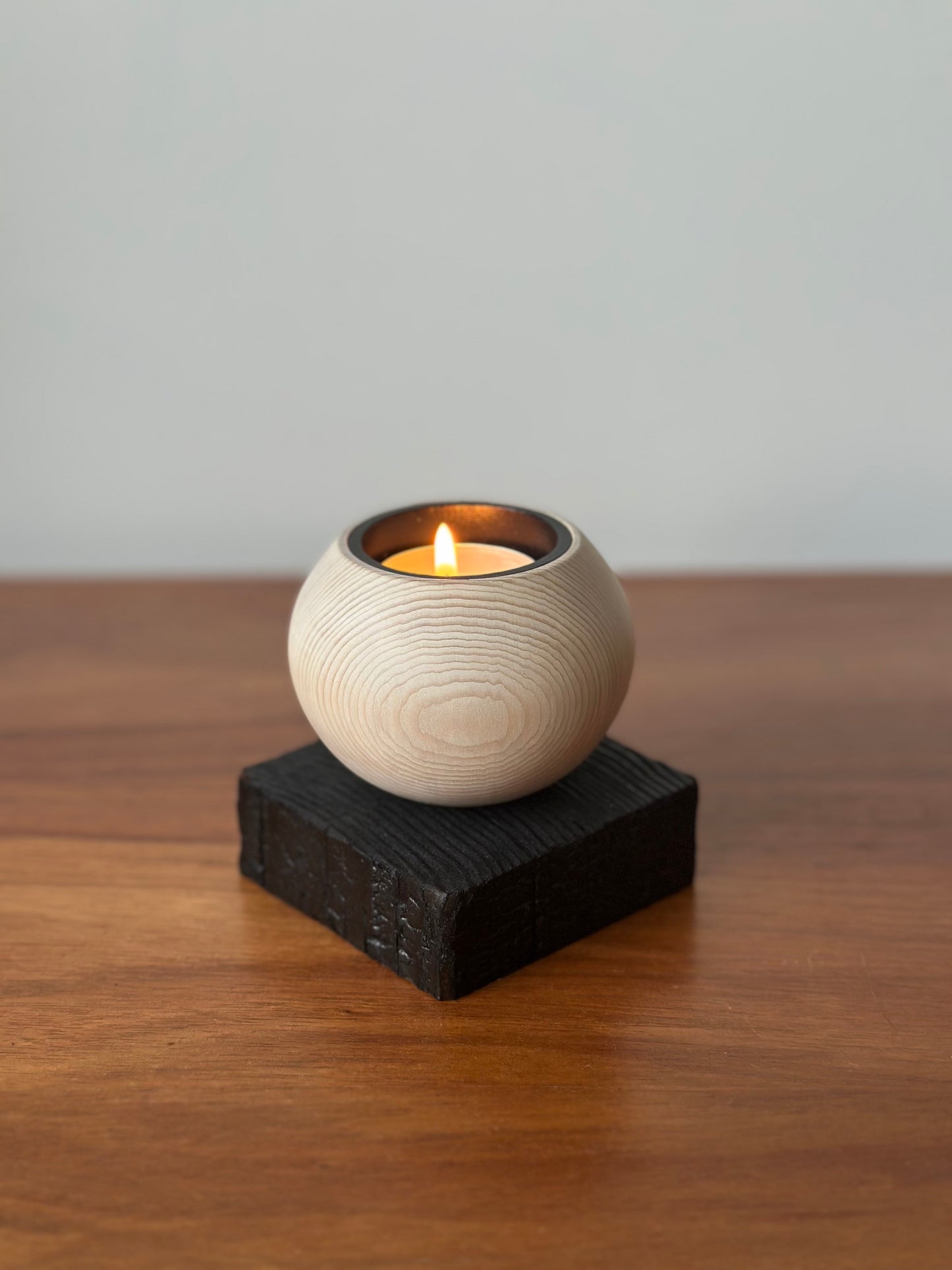 A small lit candle in a round textured cream holder, placed on a dark rectangular block atop a wooden tabletop against a plain light gray wall.