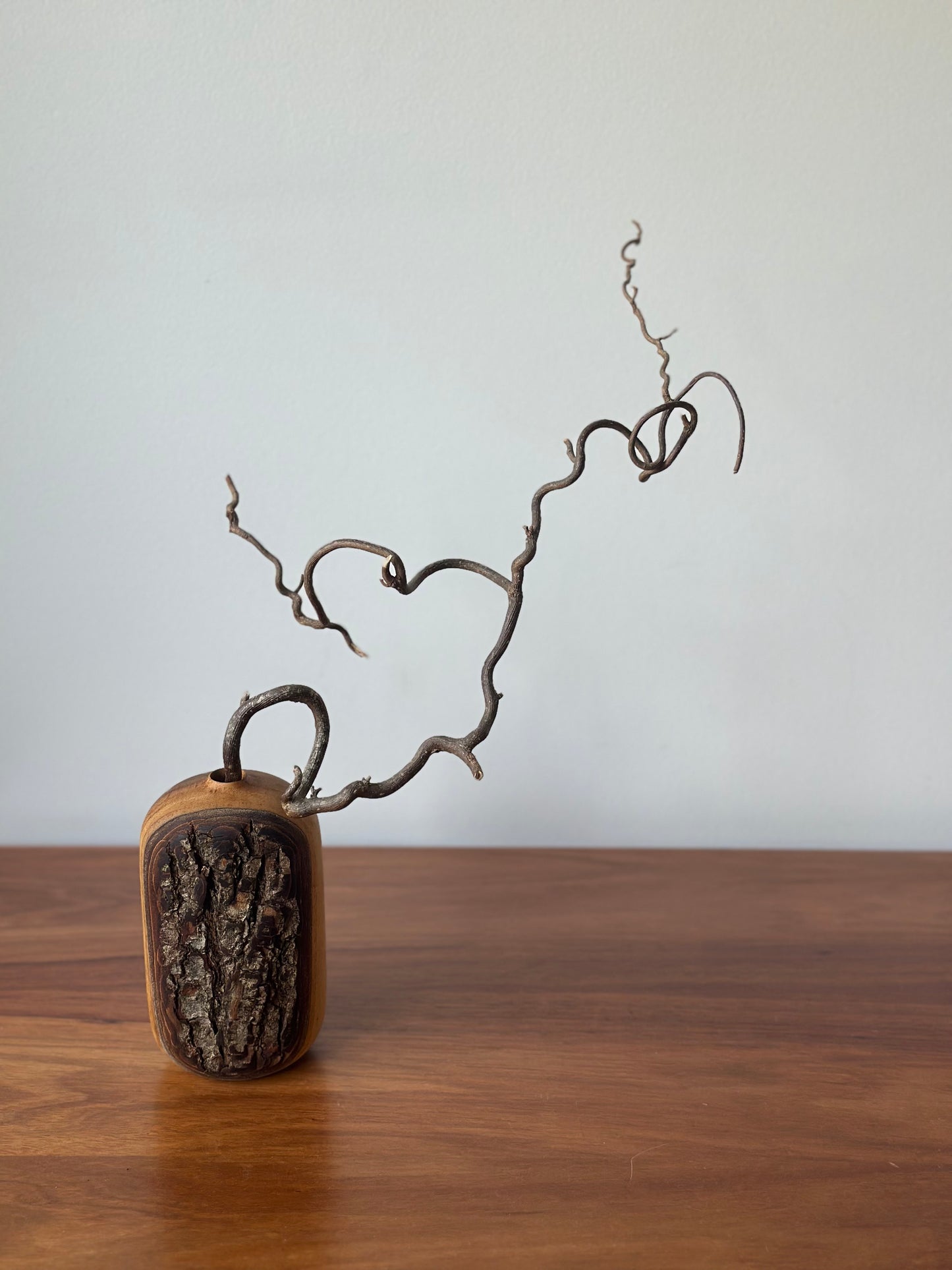 A small decorative sculpture or vase-like object with a rounded rectangular base, featuring a dark textured front panel and a curved brown branch or vine rising from the top, displayed on a wooden surface against a plain light wall.