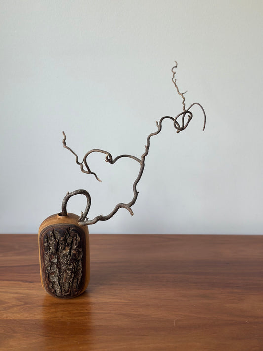 A small decorative sculpture or vase-like object with a rounded rectangular base, featuring a dark textured front panel and a curved brown branch or vine rising from the top, displayed on a wooden surface against a plain light wall.